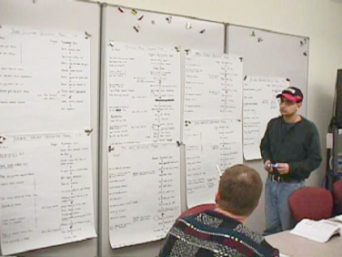 A photograph of 2 students, one is at the wall holding a pen, the other is sitting, talking and pointing at the sequence models hung by magnets onto the wall. 